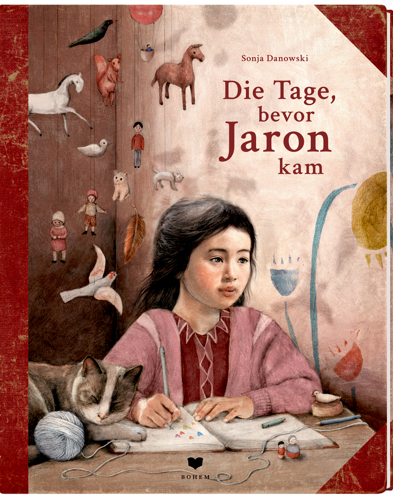 Ein Mädchen schreibt am Schreibtisch mit einer Katze und Spielzeug unter dem Titel „Die Tage, bevor Jaron kam“.