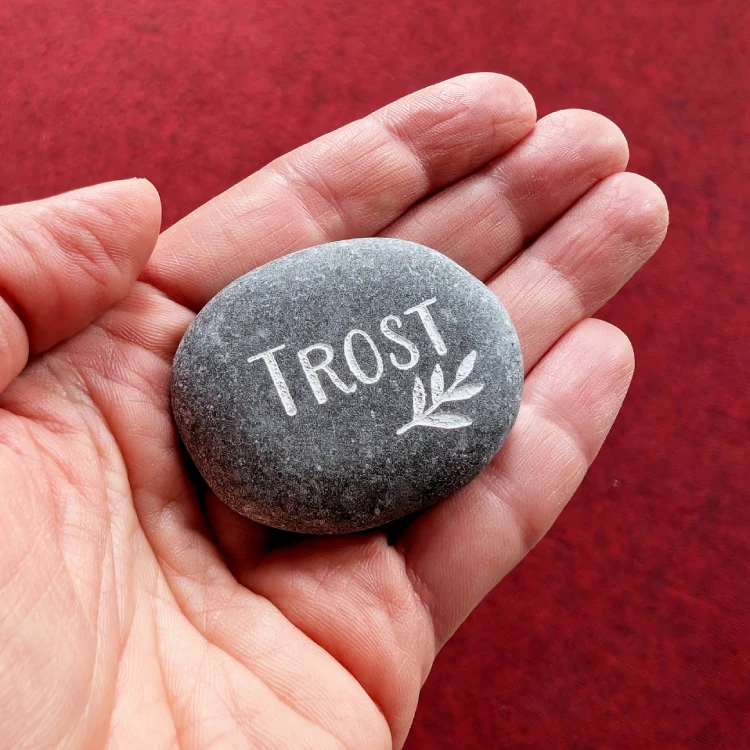 Eine Hand hält einen grauen Stein, in den das Wort "Trost" und ein Zweig eingraviert sind.
