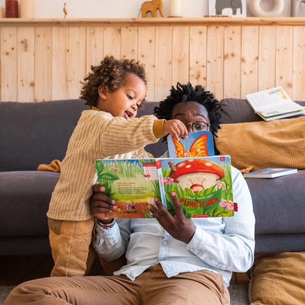 Vater sitzt angelehnt an einem Sofa und liest mit seinem Sohn gemeinsam ein Buch. Dieser steht neben ihm und zeigt etwas.