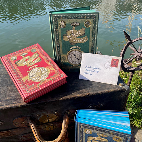 Die Coppenrath Jules Verne Schmuckausgaben stehen auf einem Koffer an einem See, an einem der Bücher lehnt ein Brief.