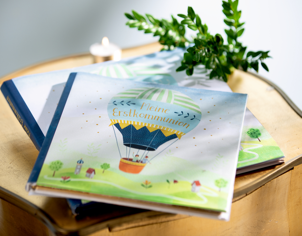 Eintragalbum „Meine Erstkommunion“ mit Heißluftballon-Illustration liegt auf einem Holztisch.