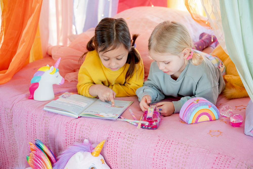 Zwei Kinder liegen auf einem rosa Bett, vor ihnen liegen ein Buch, Stifte und ein Klebebandabroller.