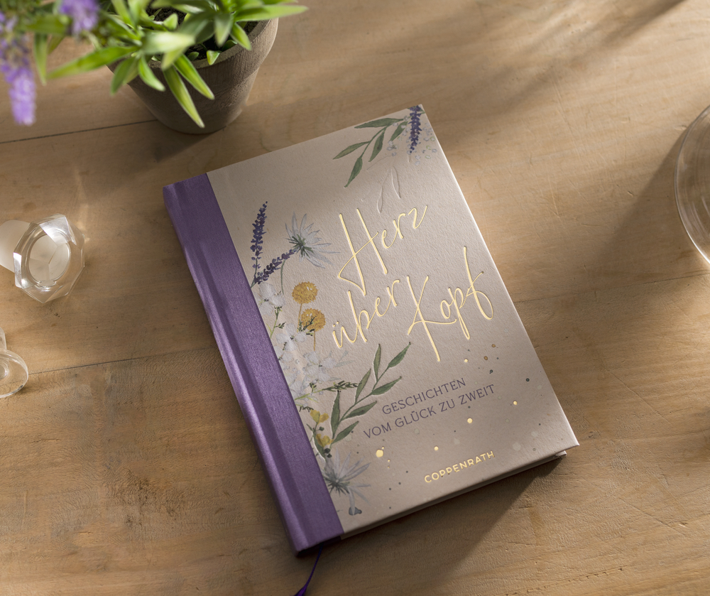 „Herz über Kopf“, ein Buch mit geblümtem Einband, liegt auf einem Holztisch neben einer Pflanze und einer Kerze.