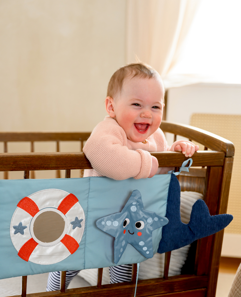 Baby im Gitterbett mit blauem Stoffbuch mit Rettungsring, Seestern und Walflosse am Bettrand.