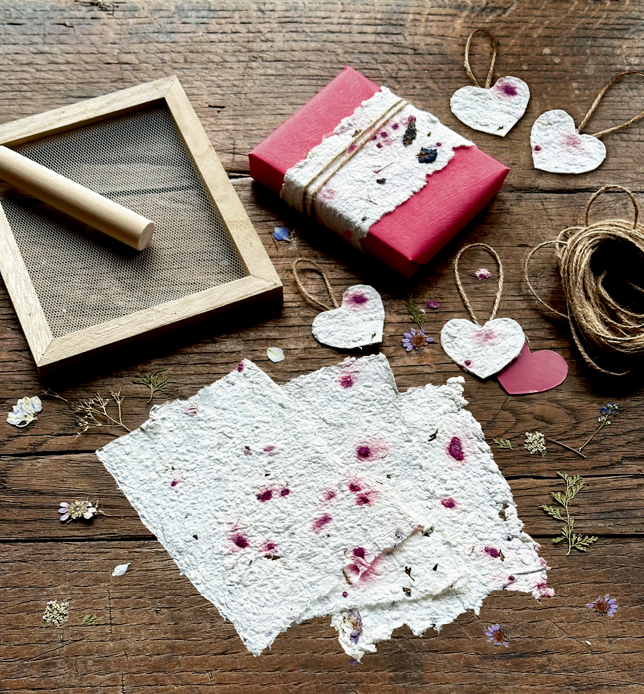 Handgeschöpftes Papier und herzförmige Tags mit gepressten Blumen liegen neben einer rosa Geschenkbox und Bastelwerkzeugen auf Holz.