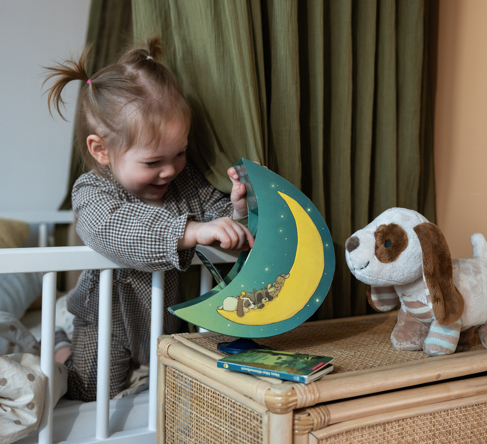 Kind im karierten Schlafanzug spielt im Bett mit Buchbox in Halbmond-Form neben einem Plüschhund.