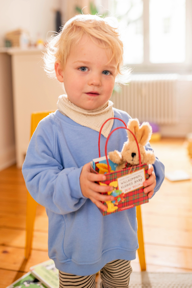 Ein Kleinkind in einem blauen Pullover steht drinnen und hält eine kleine Geschenktüte mit einem ausgestopften Hasen darin.