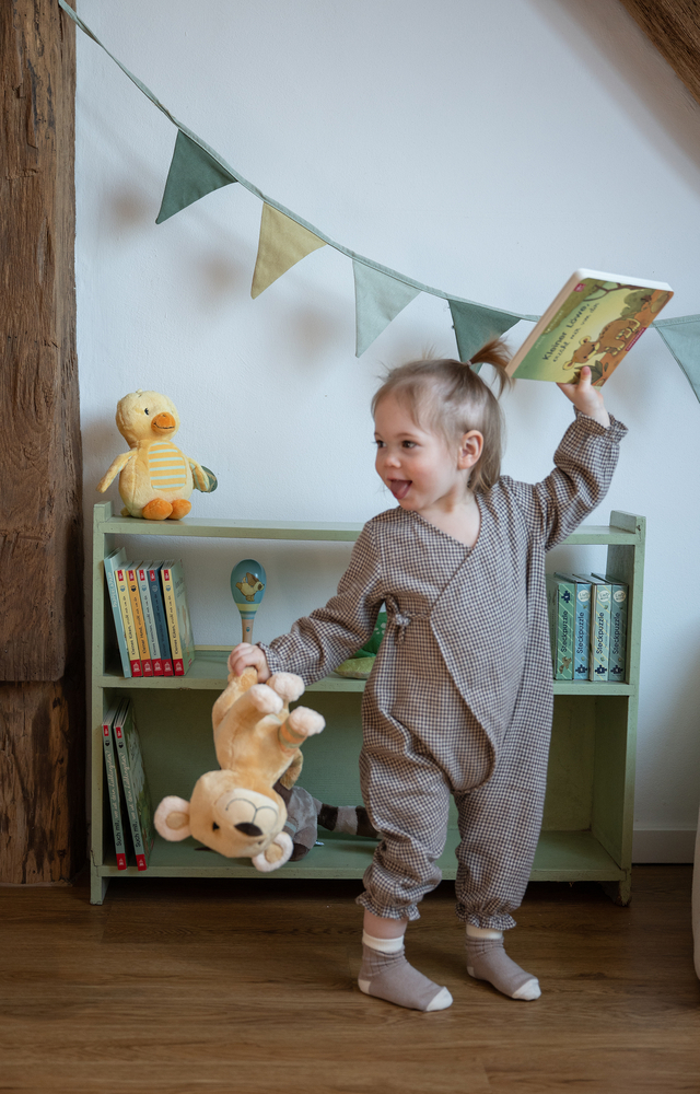 Kind hält ein Buch und ein Kuscheltier vor einem grünen Regal mit Kinderbüchern und Stofftier.