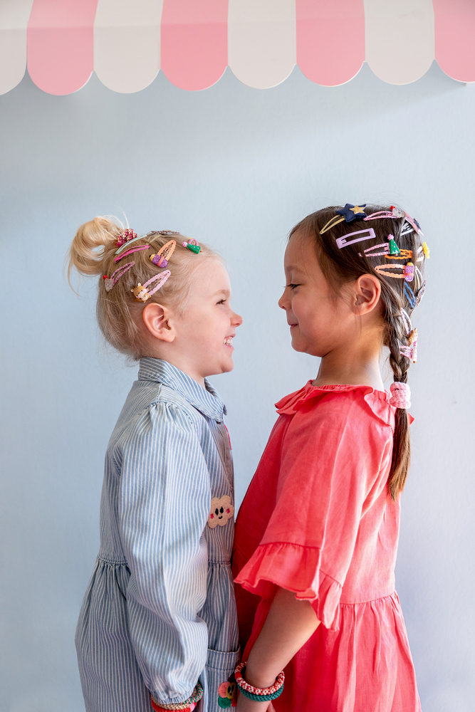 Zwei Kinder stehen sich gegenüber, tragen bunte Haarklammern und Armbänder, eines im blauen, eines im roten Kleid.