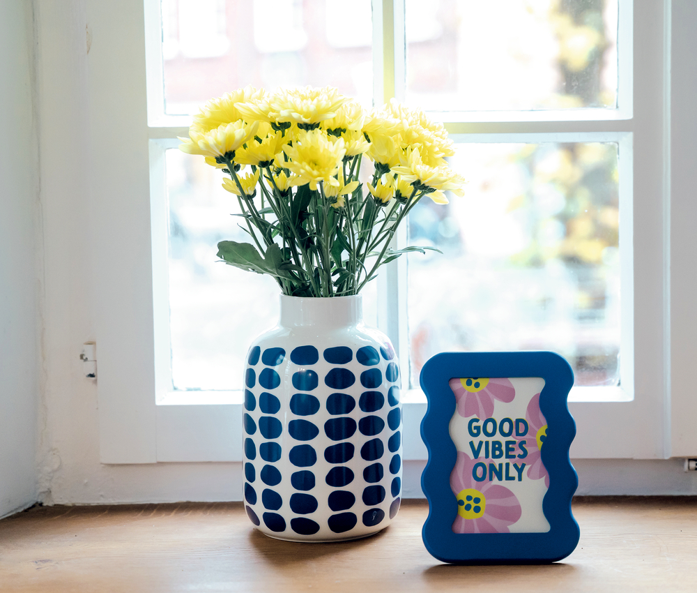 Gelbe Blumen in Vase mit blauem Muster. Daneben blauer Rahmen mit Aufschrift GOOD VIBES ONLY auf Fensterbank.