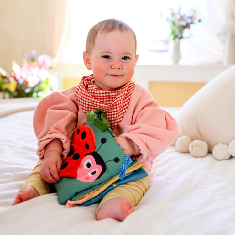 Lächelndes Baby mit buntem Stoffbuch sitzt auf einem Bett.