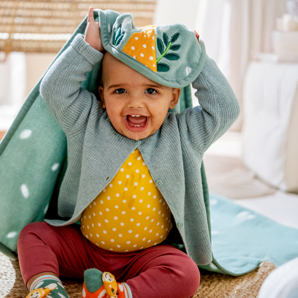 Lachendes Baby zieht sich eine türkis-grüne Decke über den Kopf.