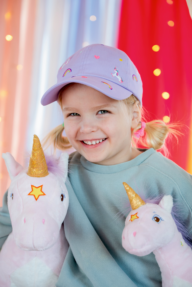 Kind mit lila Kappe mit Einhorn- und Regenbogenmotiven hält zwei rosa Einhorn-Plüschtiere mit goldenen Hörnern.