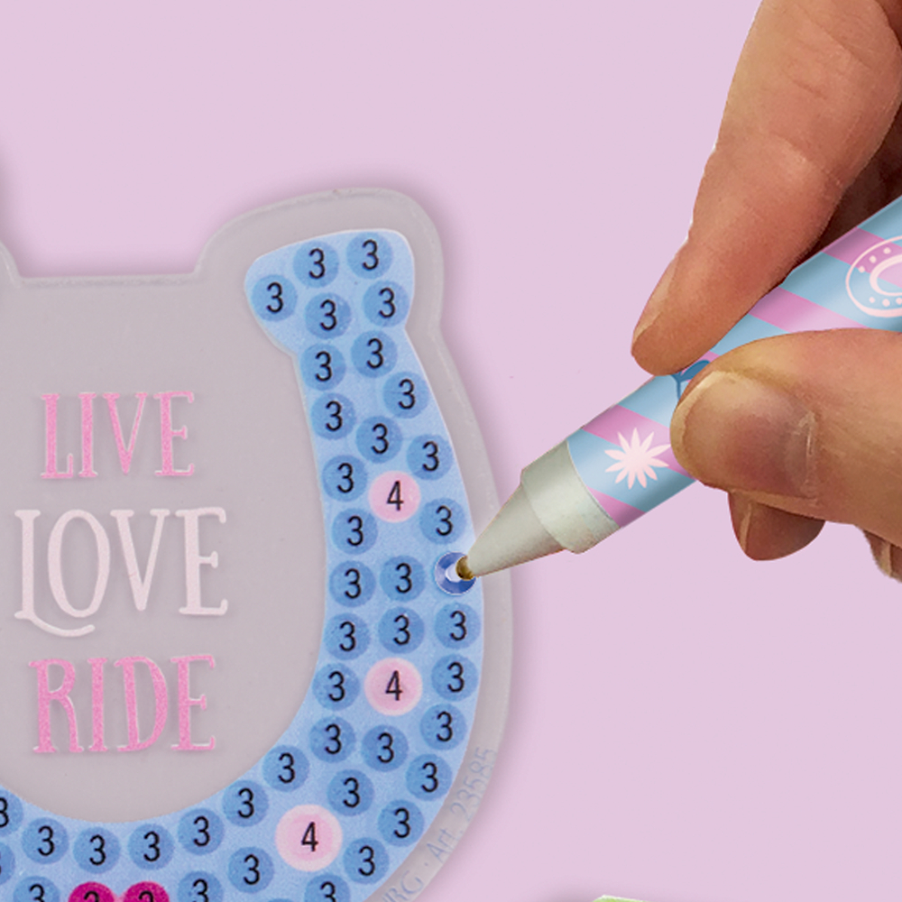 Hand trägt mit Stift blaue Steinchen auf nummerierte Fläche in Hufeisenform mit rosa Schrift „LIVE LOVE RIDE“ auf.