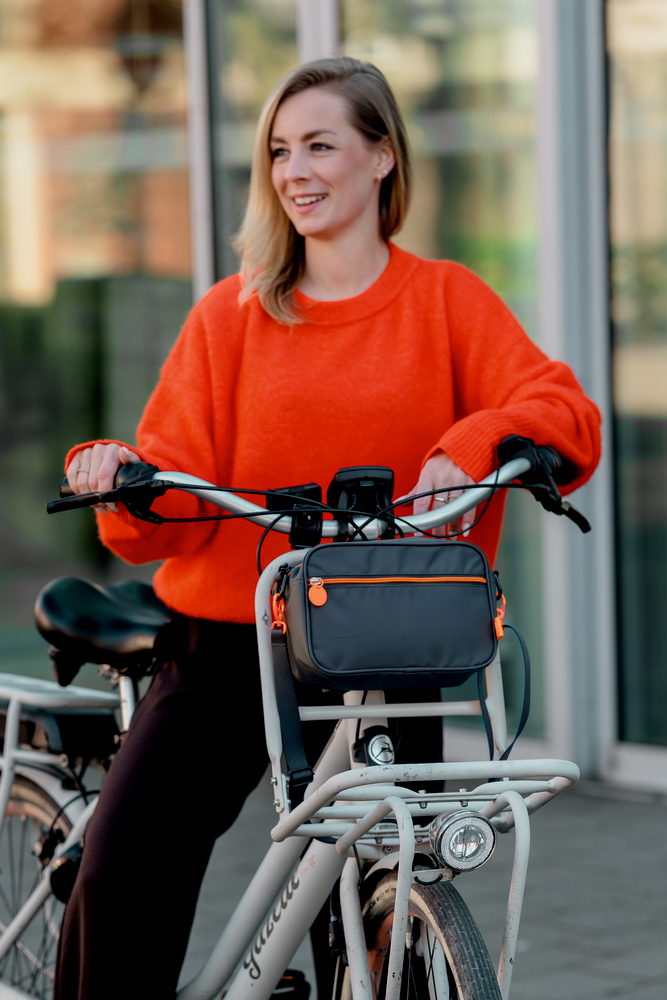 Person in orangefarbenem Pullover steht mit einem hellgrauen Fahrrad mit grauer Lenkertasche mit orangefarbenen Details.