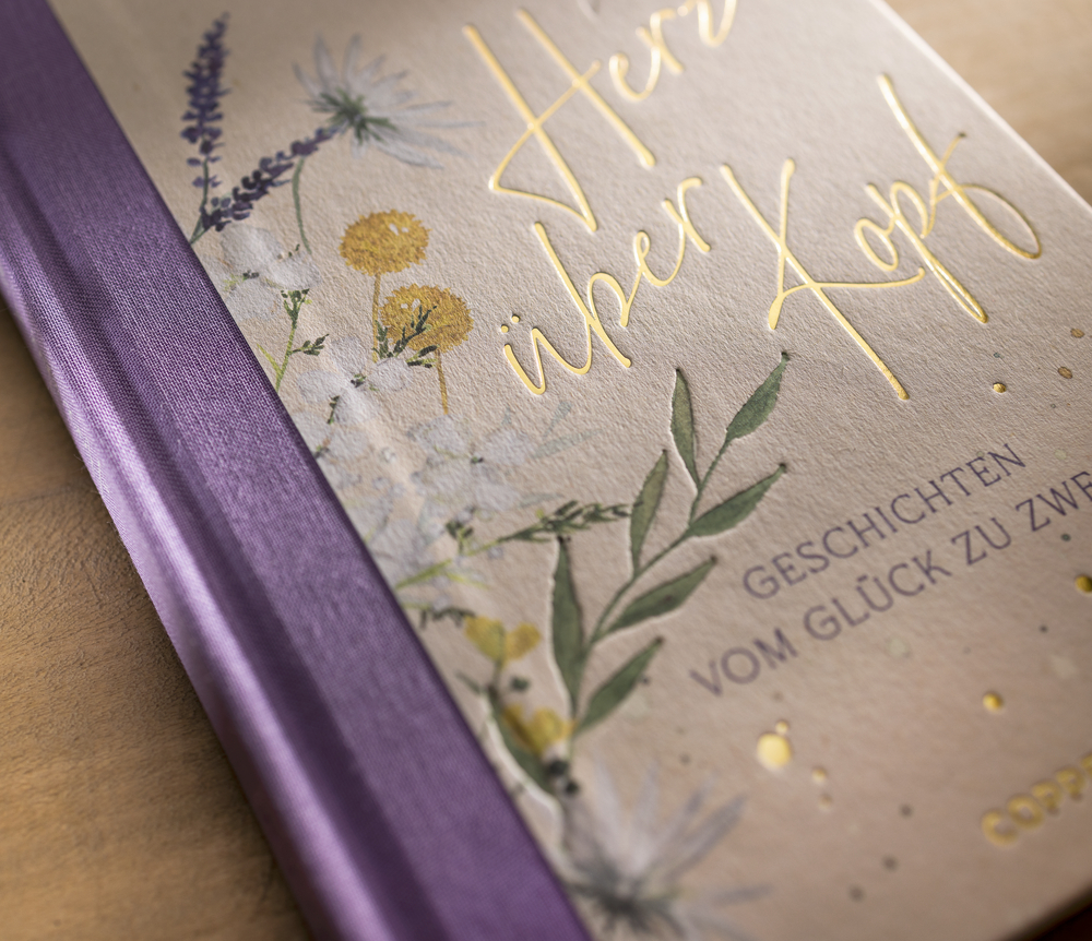 Ein Buch mit violettem Buchrücken, floralem Coverdesign und dem goldenen Text „Herz über Kopf“ auf der Vorderseite.