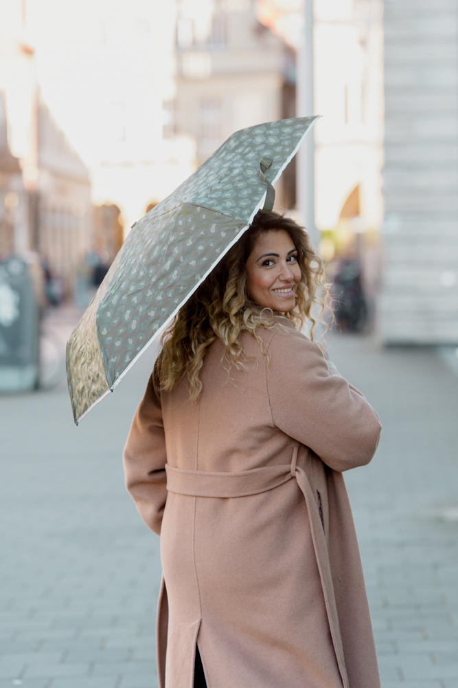 Person in hellbraunem Mantel hält einen Regenschirm mit hellem Muster auf einer Straße.