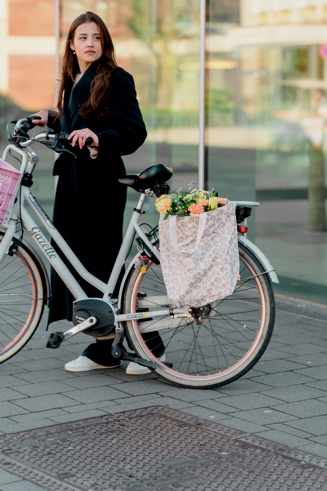 Person in dunklem Mantel mit grauem Fahrrad, an dessen Gepäckträger eine Fahrradtasche gefüllt mit Blumen hängt.