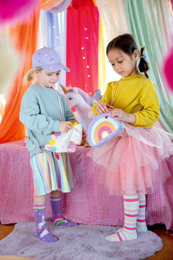 Zwei Kinder mit bunten Taschen im Einhorn- und Regenbogendesign in farbig dekoriertem Raum.