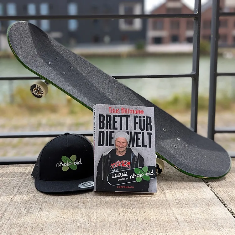 Ein Skateboard lehnt schräg an einem Metallgeländer. Vor dem Skateboard steht Titus Buch "Brett für die Welt" und eine Skate-Aid Kappe