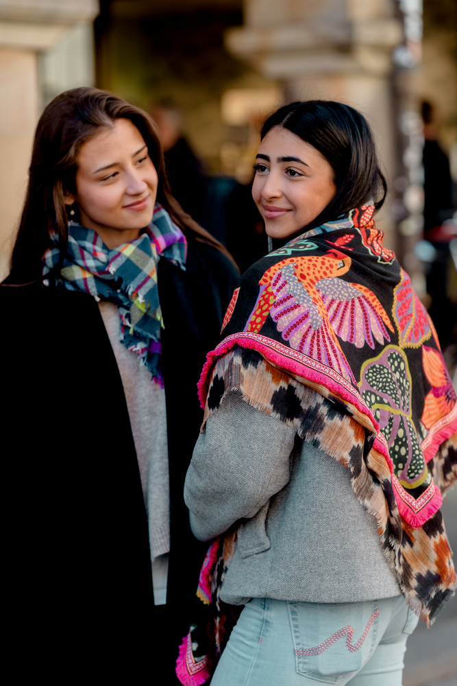 Zwei Personen mit bunten Schals stehen nebeneinander, eine trägt eine graue Jacke und darüber eine bestickte bunte Schulterstola.