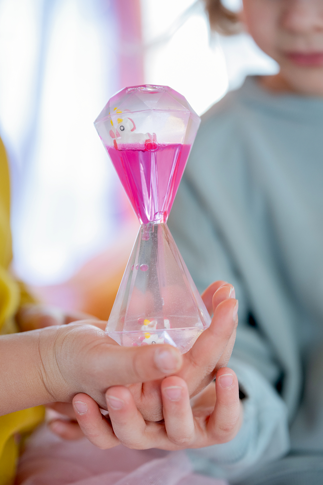 Kind hält eine transparente, diamantförmige Sanduhr mit rosa Flüssigkeit und kleinem Einhornmotiv.