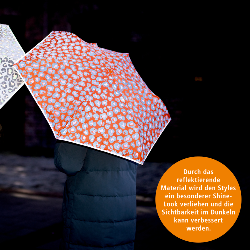 Person mit dunkelblauer Jacke hält orangenen Regenschirm mit reflektierendem Leomuster bei Dunkelheit.