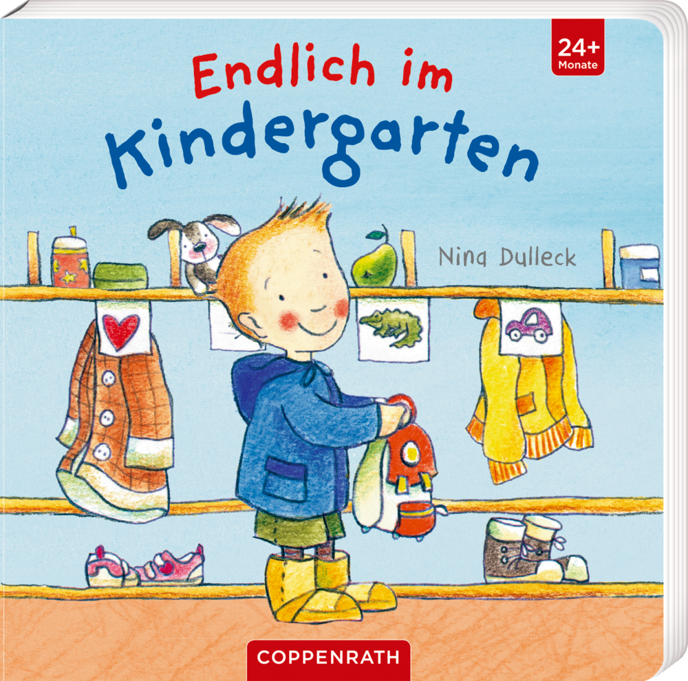 Ein lächelndes Kind in einem blauen Mantel steht neben den Ablagefächern in der Garderobe eines Kindergartens, auf dem Cover von "Endlich im Kindergarten".