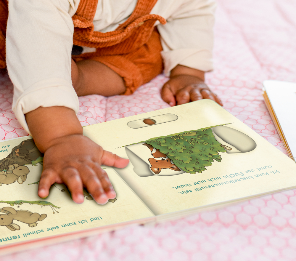 Ein Baby im orangefarbenen Overall berührt ein Bilderbuch, während es auf einer rosa Decke liegt.