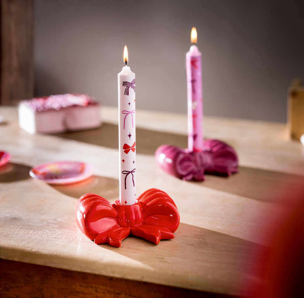 Zwei brennende Stabkerzen mit Schleifenmuster in roten und pinken Kerzenhaltern in Schleifenform auf einem Holztisch.