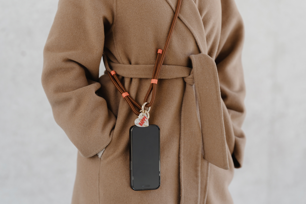 Smartphone an braun-orangefarbener Kordel mit Anhänger und Aufschrift „HAPPY“, getragen über beigem Mantel.