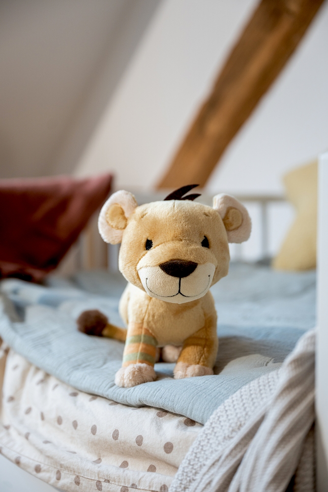 Beiger Plüschlöwe mit brauner Nase und gestreiftem Bein sitzt auf einem Kinderbett mit hellblauer Decke und Kissen.