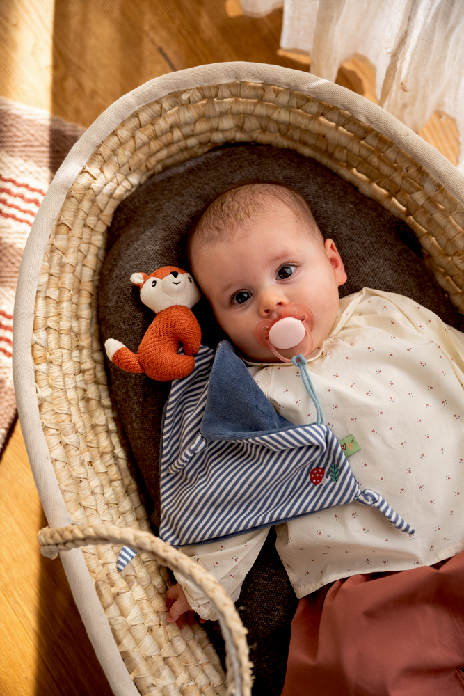 Baby mit Schnuller liegt in einem Korb auf dunkler Decke, neben ihm ein Gestreiftes Schnullertuch mit Fuchs-Stofffigur.