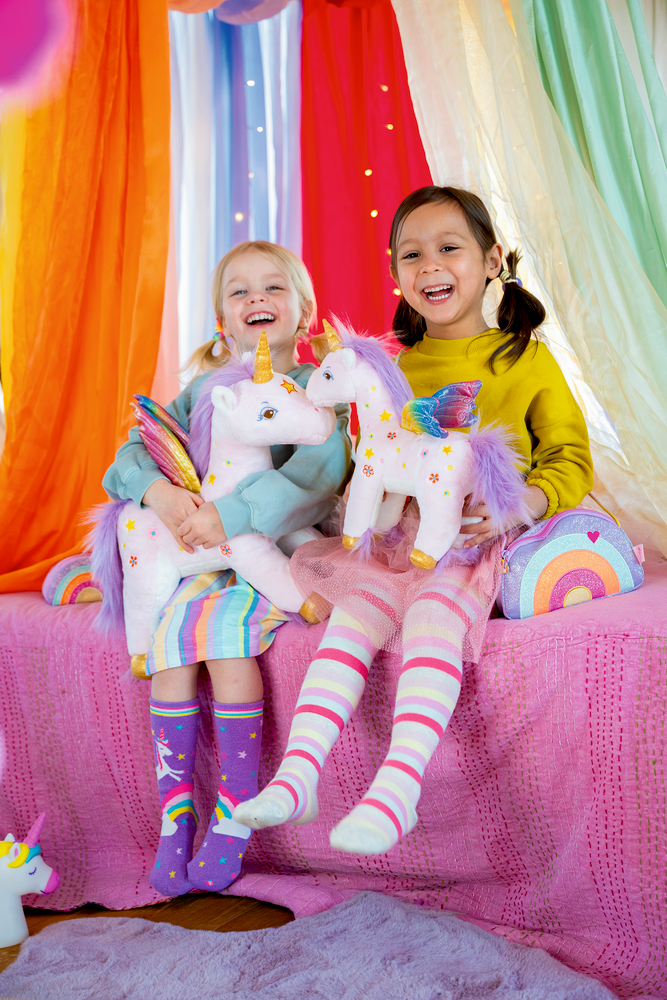 Zwei Kinder sitzen auf rosa Decke und halten weiße Einhorn-Plüschtiere mit bunten Flügeln und goldenen Hörnern.