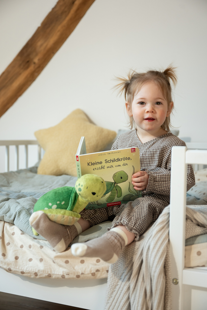 Kind in karierter Kleidung sitzt auf Bett, hält ein Kinderbuch mit Schildkrötenmotiv und hat eine Plüschschildkröte auf dem Schoß.