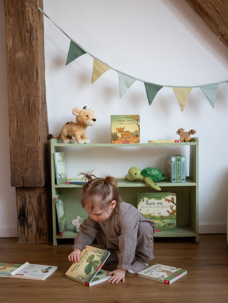 Kleines Kind sitzt auf dem Boden vor einem Regal mit Kinderbüchern und Stofftieren und blättert in einem Buch.