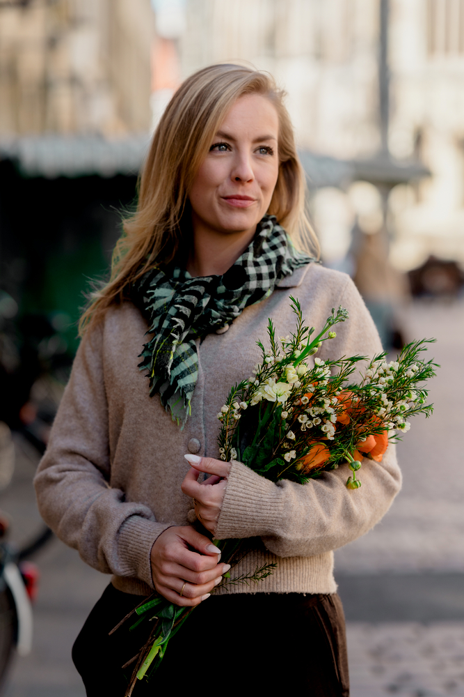 Frau mit hellbraunem Pullover und kariertem Schal hält einen Strauß aus weißen und orangefarbenen Blumen.