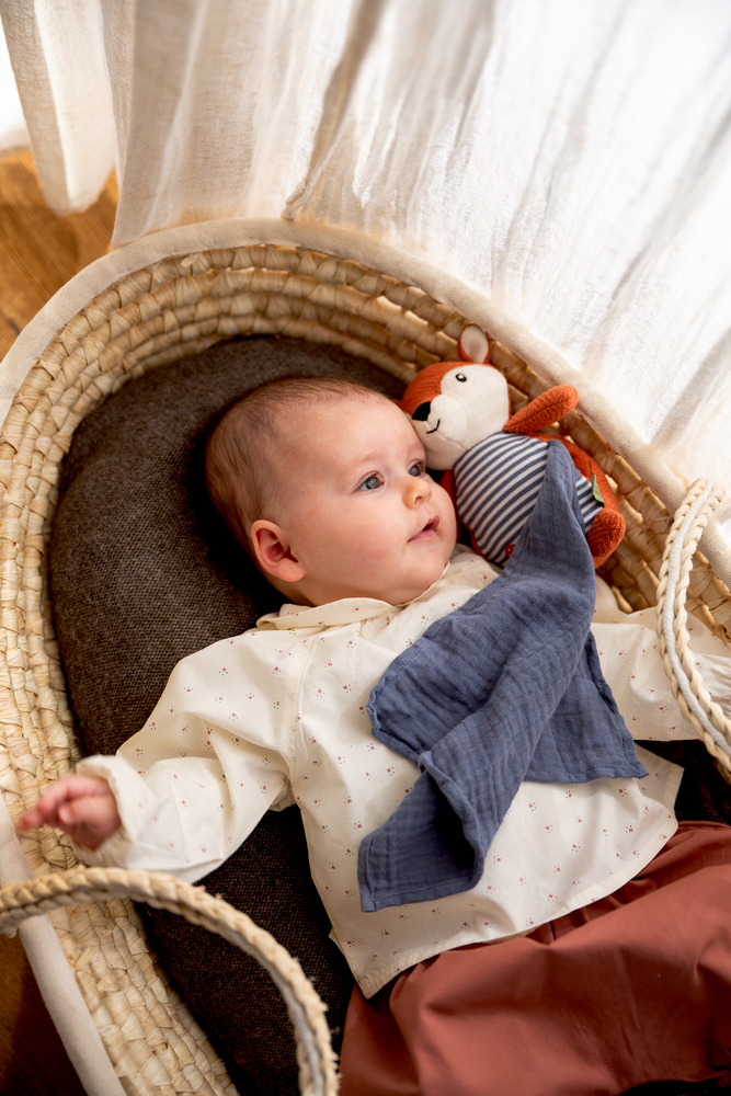 Baby liegt in einem geflochtenen Korb auf dunklem Kissen mit Stofftier und blauem Schnuffeltuch.