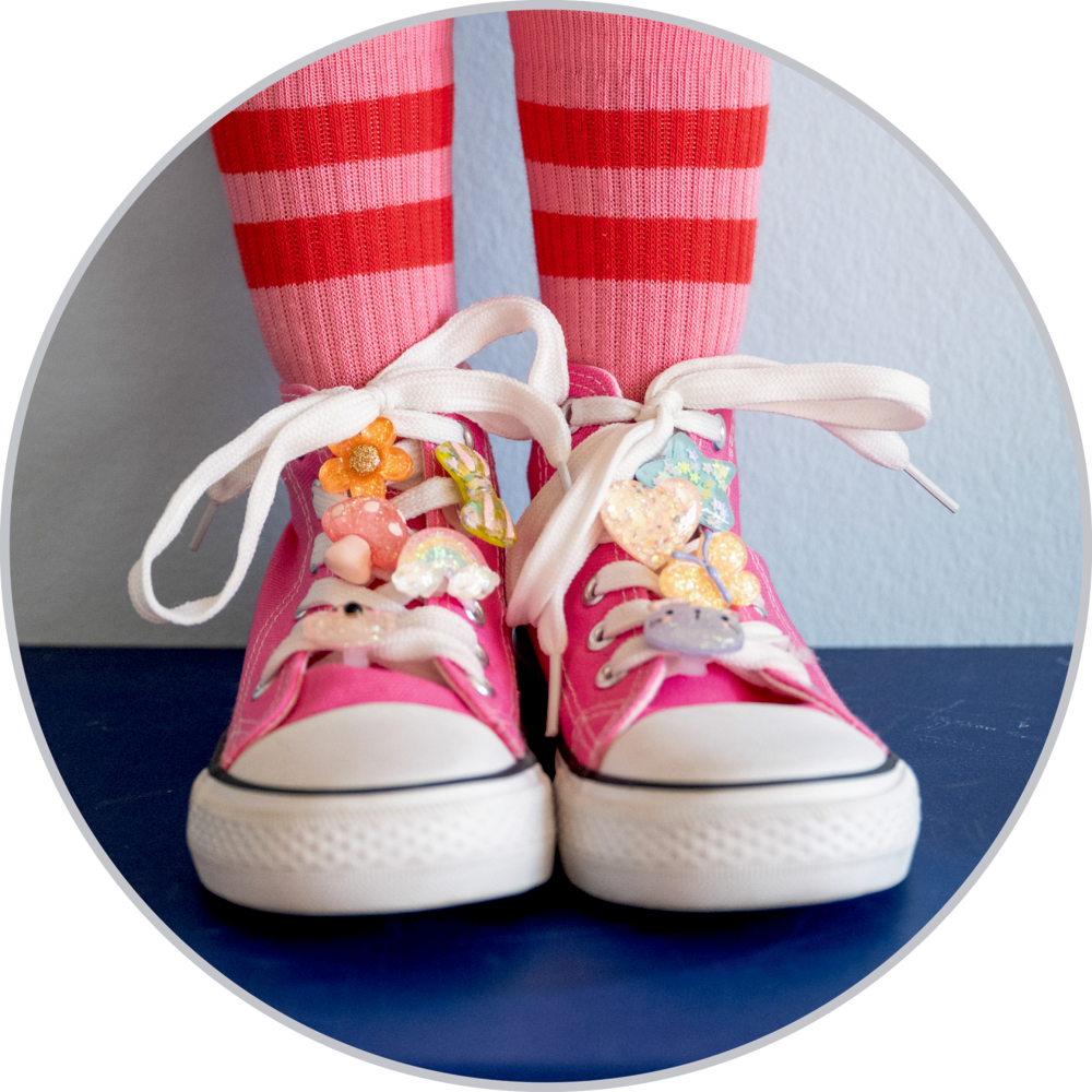 Pinkfarbene Kindersneaker mit weißen Schnürsenkeln, verziert mit bunten Sneaker Charms, dazu rosarot gestreifte Socken.