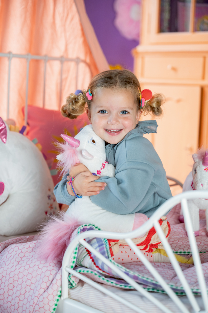 Mädchen umarmt Plüscheinhorn auf Bett in buntem Kinderzimmer.