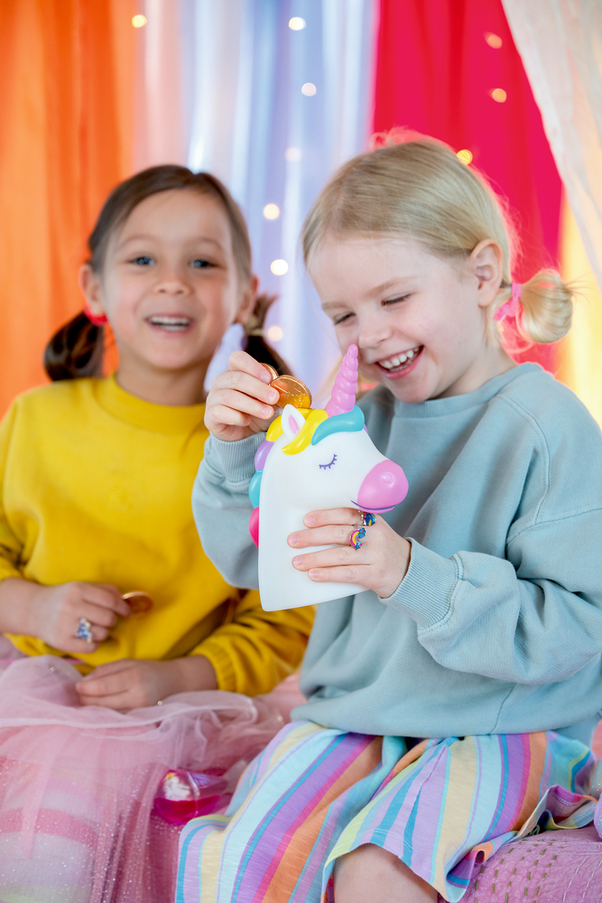 Zwei Kinder stecken Münzen in eine Einhorn-Spardose mit bunter Mähne.