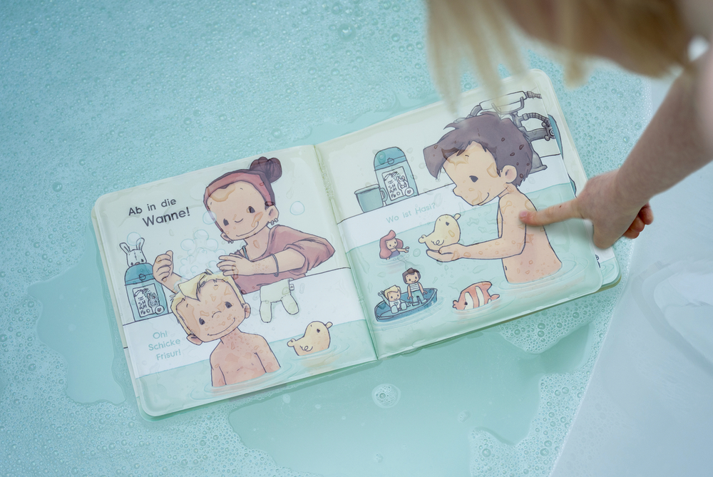 Kinderhand hält aufgeschlagenes wasserfestes Badebuch mit Illustrationen von badenden Kindern über Wasser.