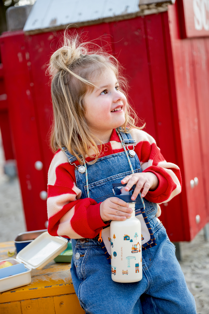 Kind hält weiße Trinkflasche mit bunten Fahrzeugmotiven und trägt rote Ringelpullover sowie blaue Latzhose vor rotem Hintergrund.