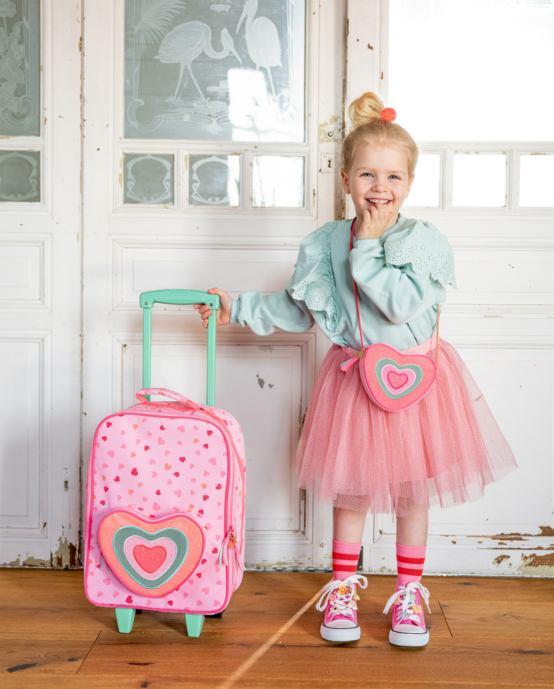 Kind mit rosa Tüllrock, herzförmiger Tasche und pinkem Rollkoffer mit Herzmotiv steht vor heller Holztür.