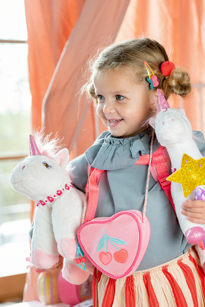 Mädchen lächelt, hält zwei Einhorn-Plüschtiere, trägt pinke Umhängetasche mit Herz und bunte Haarklammern.