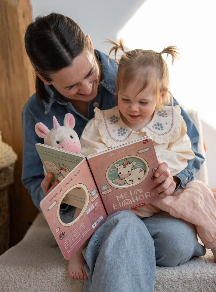Erwachsene und Kind sitzen zusammen und schauen in ein Kinderbuch mit Einhornmotiv, neben dem Buch steckt ein Einhorn-Stofftier.
