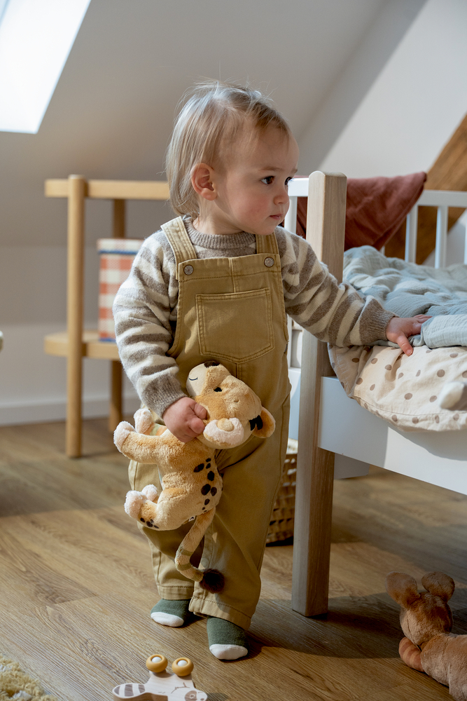 Kleinkind in beiger Latzhose und gestreiftem Pullover hält einen Plüschtier-Löwen neben einem Kinderbett.