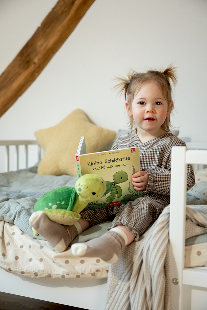 Kind auf Bett hält Kinderbuch und Plüschschildkröte, im Hintergrund Kissen und Decke.