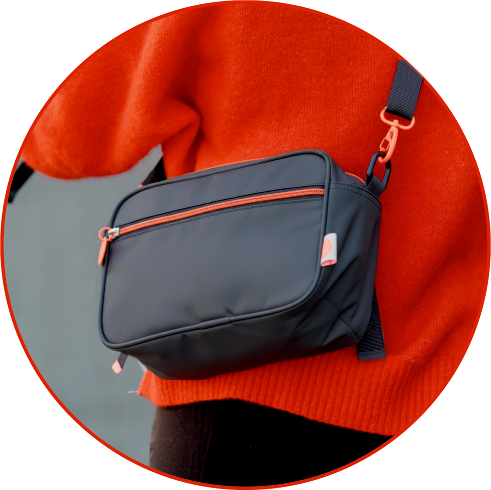 Dunkelgraue Umhängetasche mit orangenen Reißverschlüssen und Details, getragen über einem orangefarbenen Pullover.